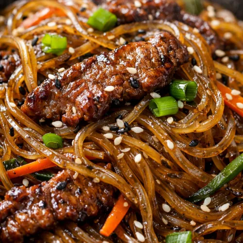 A close-up shot of steaming Mongolian Beef Glass Noodles in a dark bowl, showing glossy sweet potato noodles, seared beef pieces, green onions, and sesame seeds.