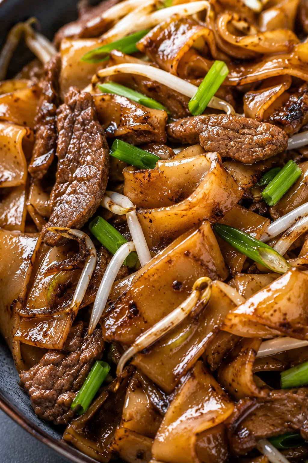 Extreme close-up of glossy Cantonese chow fun noodles stir-fried with tender beef slices, bean sprouts, and green onions in a rich soy-based sauce with visible wok-char texture and steam rising.